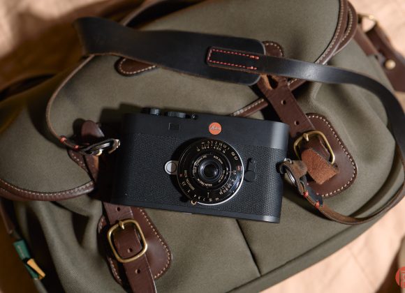 Black Leica camera resting on a khaki and brown leather strap bag, photographed from above.