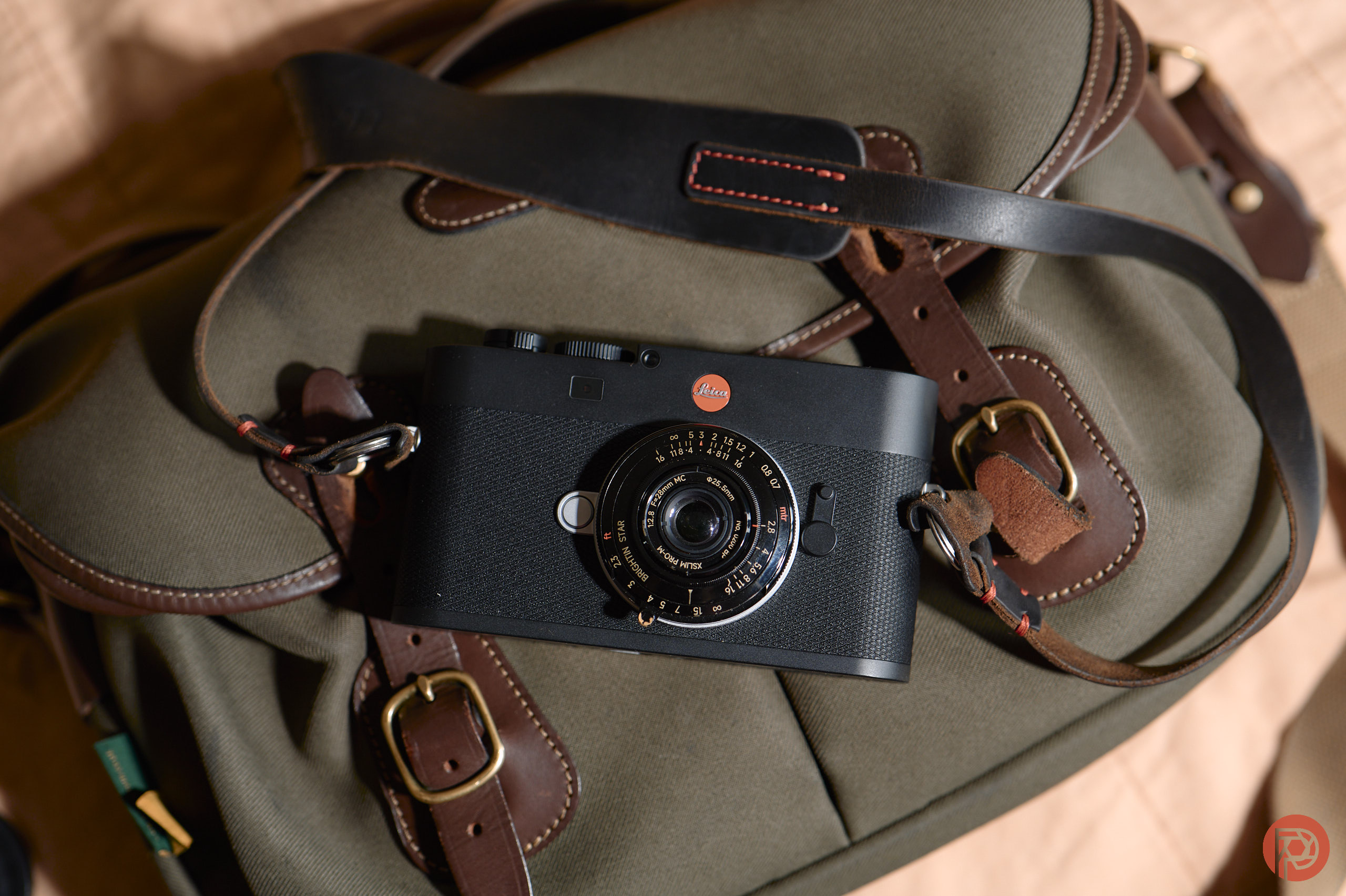 Black Leica camera resting on a khaki and brown leather strap bag, photographed from above.