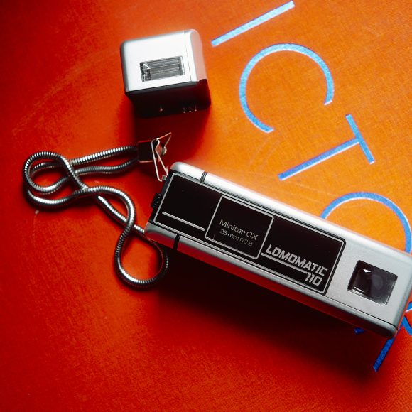 A vintage silver Lomomatic 110 camera with flash on an orange surface with blue lettering.