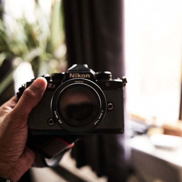 A hand holding a Nikon film camera in front of a blurred indoor background with plants and a window.