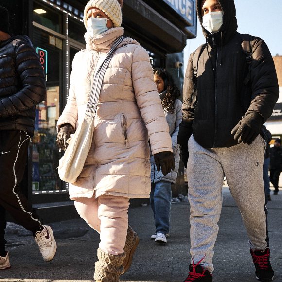 People wearing winter clothing and masks walking past a convenience store on a sunny day.