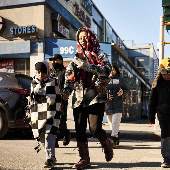 People cross a city street on a sunny day, bundled up in winter clothes and blankets.