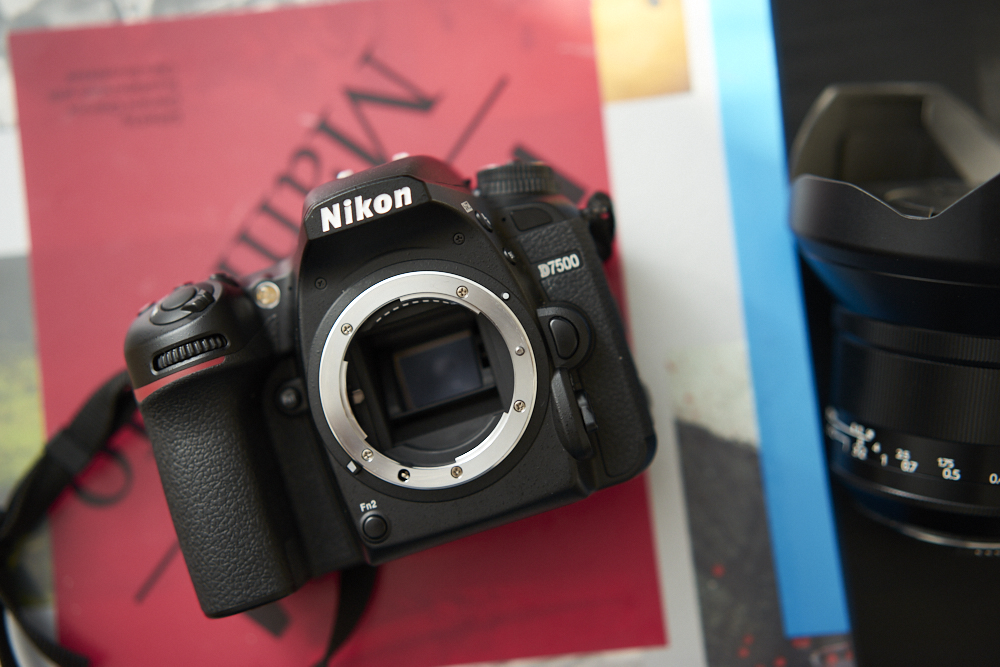 A Nikon D750 camera body is placed on a red magazine next to a detached camera lens.