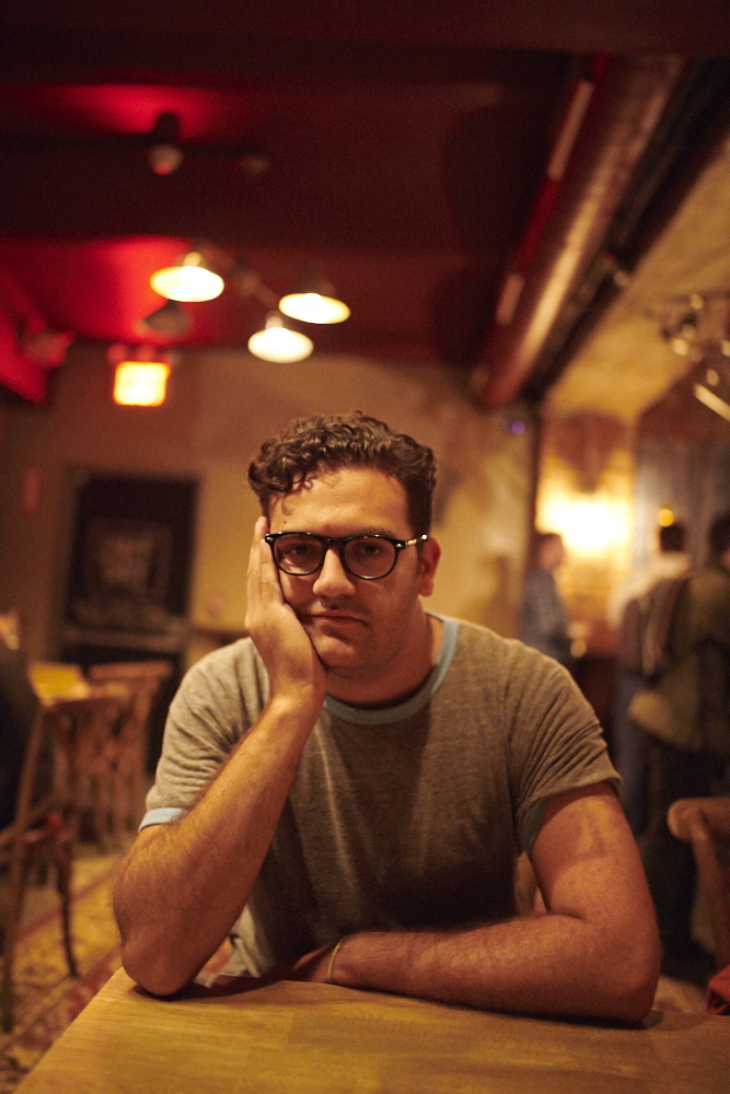 A man with glasses sits at a table, resting his face on his hand in a dimly lit café or bar.