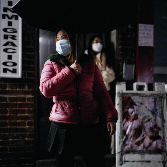 Woman in a pink jacket and mask holds a purple umbrella at night near an Immigracion sign.