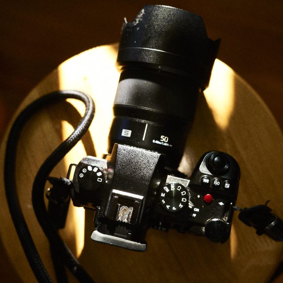 A Panasonic S5 II camera with a 50mm lens rests on a round wooden surface. The cameras numerous dials, buttons, and top-viewfinder are prominently visible. A strap is attached to the sleek design, while the lighting artfully casts shadows across the wood.
