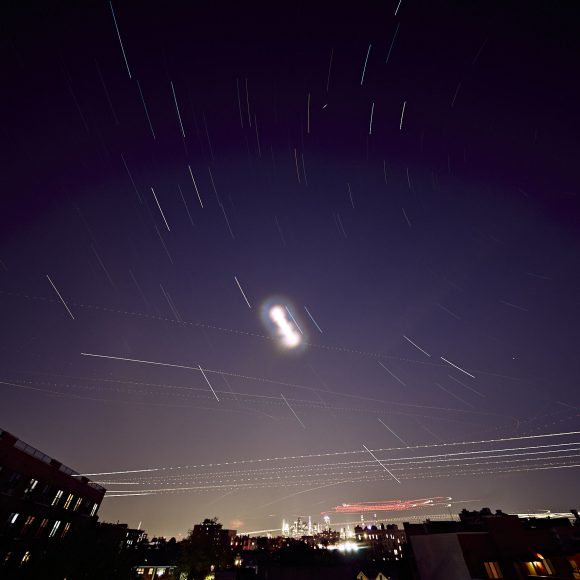 Long exposure photo of city skyline at night showing star trails and light streaks from airplanes in the sky.