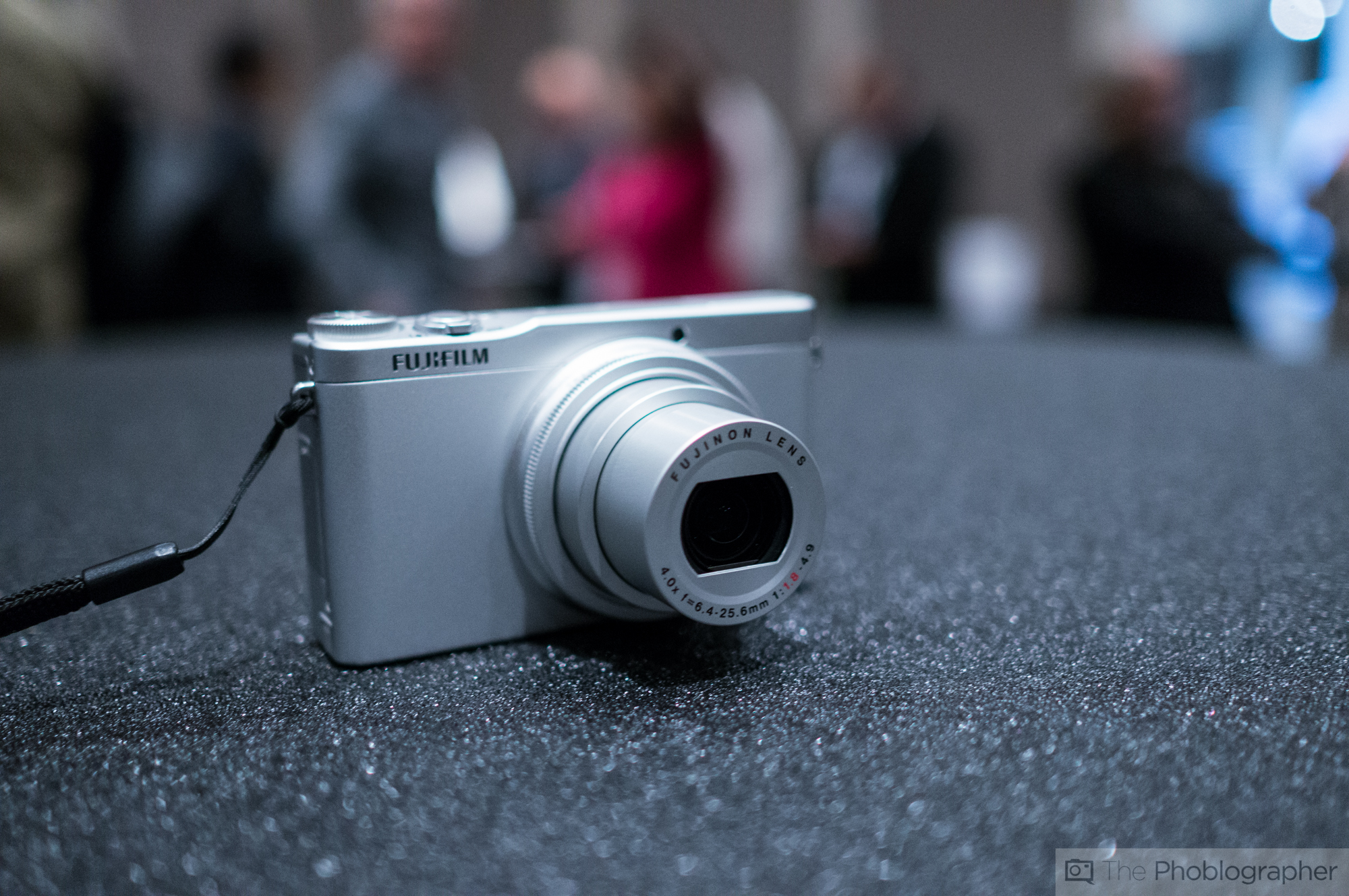 A silver Fujifilm digital camera is displayed on a textured black surface with a blurred background.