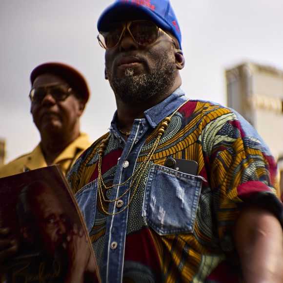 Two men stand outside; one in a colorful shirt and blue cap holds a vinyl record, the other wears glasses and a beret.