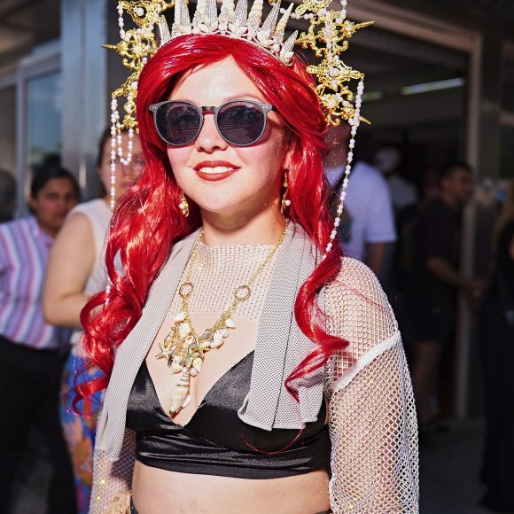 Person with long red hair, sunglasses, ornate gold crown, and stylish festival outfit standing outdoors.