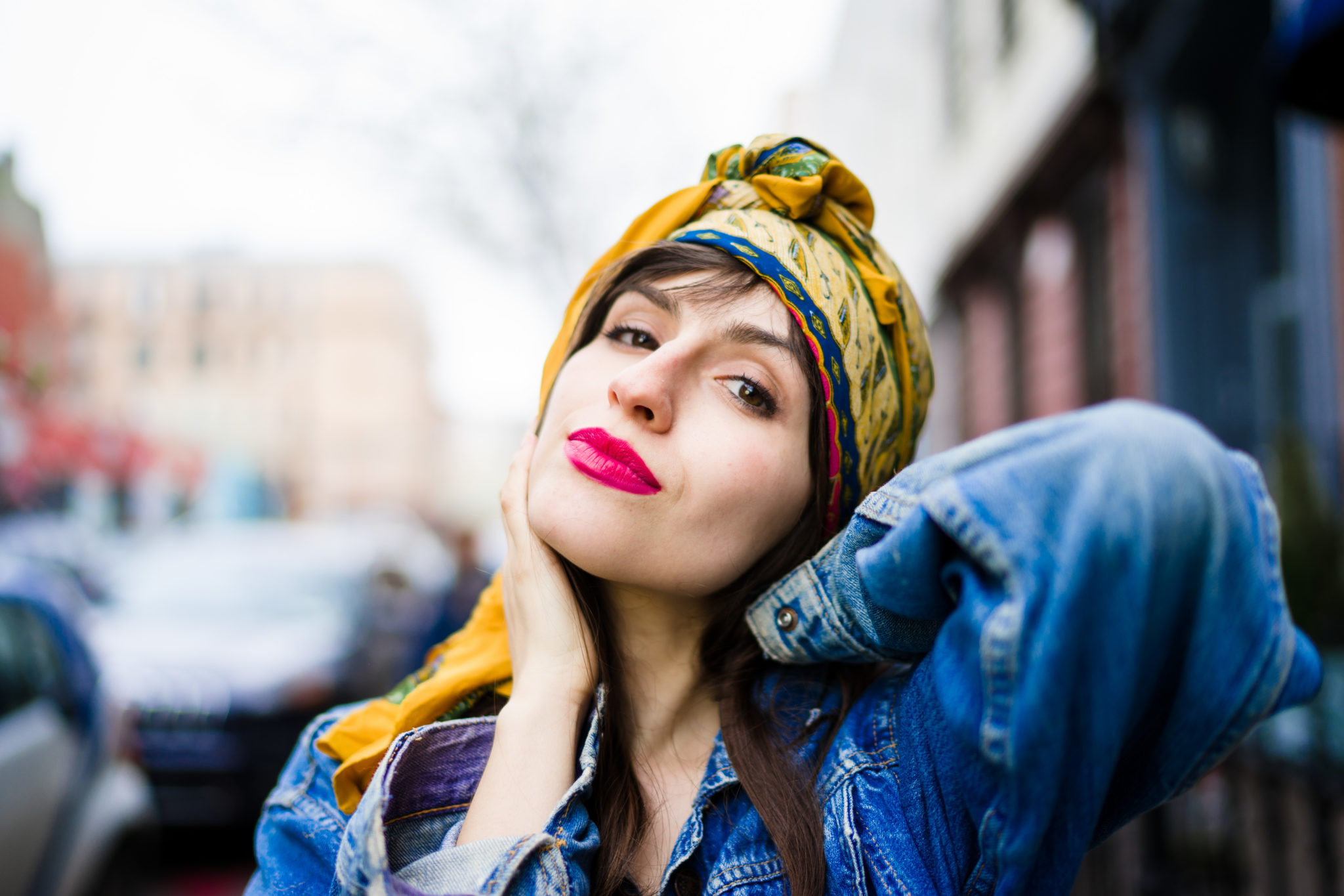 Woman wearing a yellow headscarf and denim jacket, posing confidently outdoors with one hand on her face.
