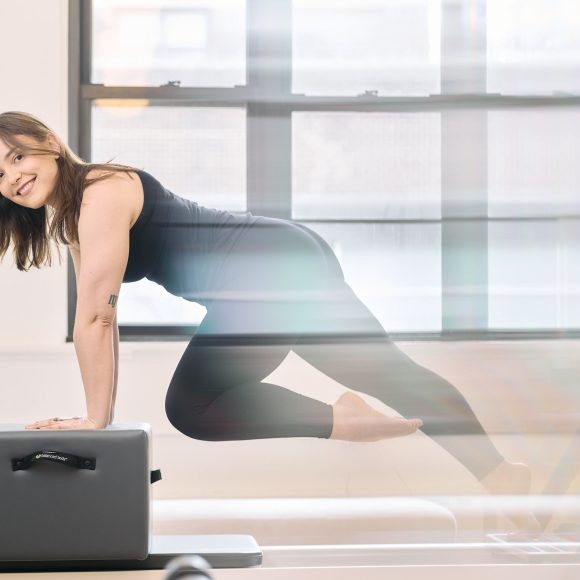 Woman doing Pilates, balancing on a reformer in a well-lit studio. She is wearing black athletic wear and smiling.