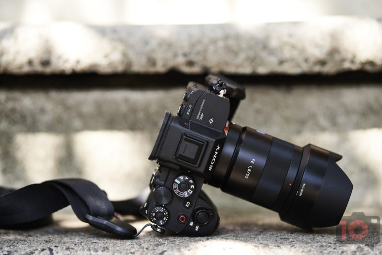 A black Sony camera with a zoom lens rests sideways on a stone surface in natural light.