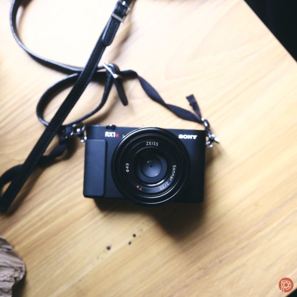 A black Sony RX1 camera with a Zeiss lens sits on a light wooden surface with its strap attached.