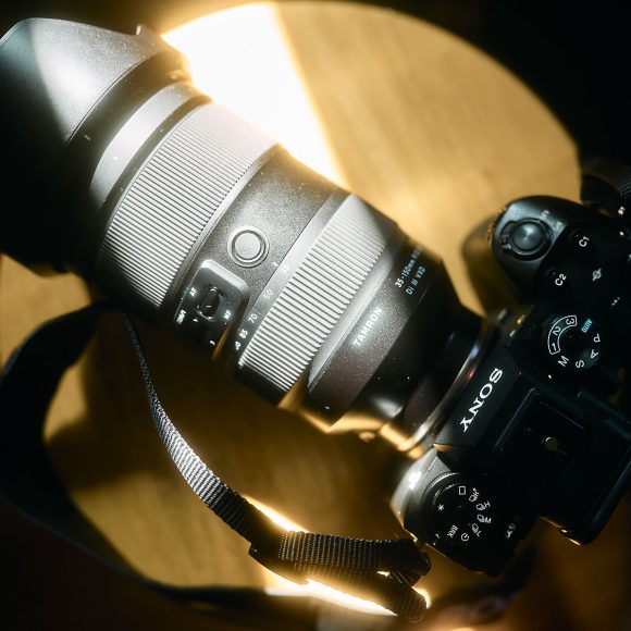 A Sony camera with a large zoom lens rests on a wooden surface, illuminated by sunlight.