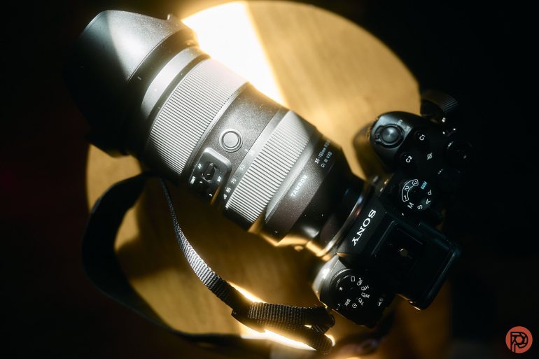 A Sony camera with a large zoom lens rests on a wooden surface, illuminated by sunlight.