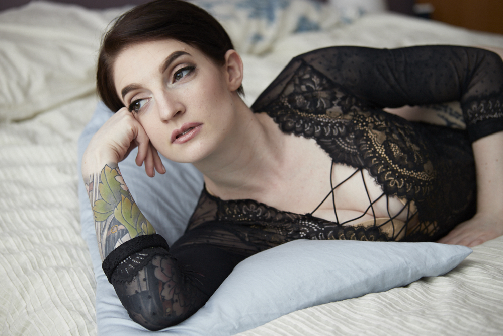 A woman in black lace clothing lies on a bed, resting her head on a blue pillow and looking to the side.