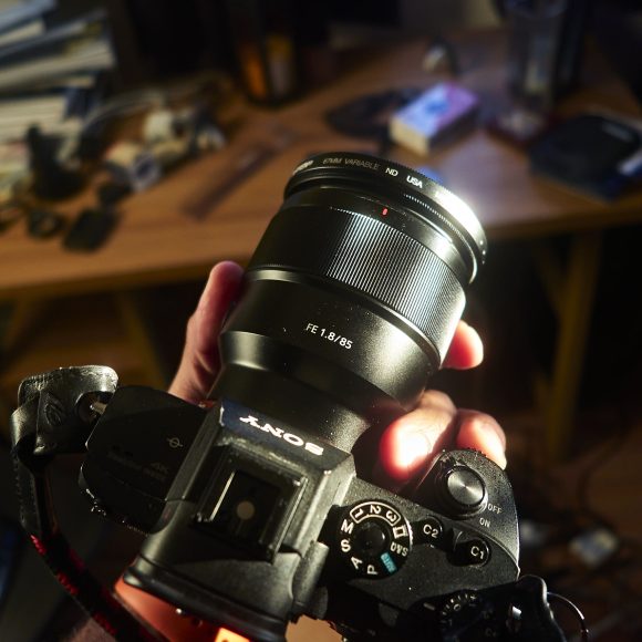 Close-up of a person holding a Sony camera with a large lens, desk and clutter visible in the background.