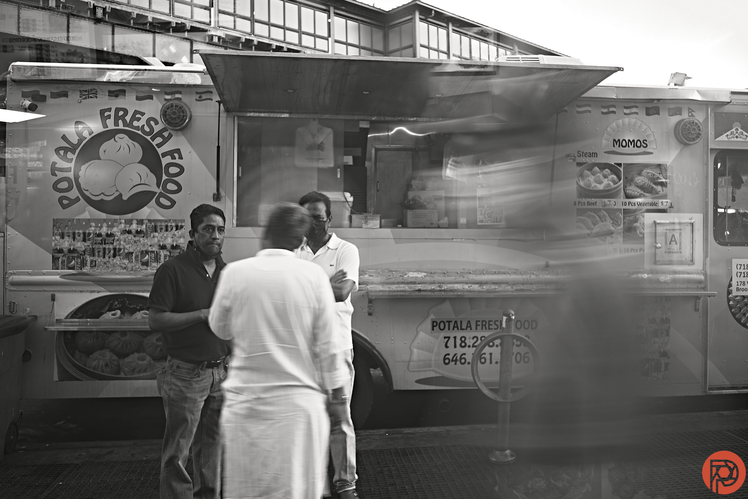 Three men stand talking in front of a Potala Fresh Food truck, with a blurred passerby in the foreground.