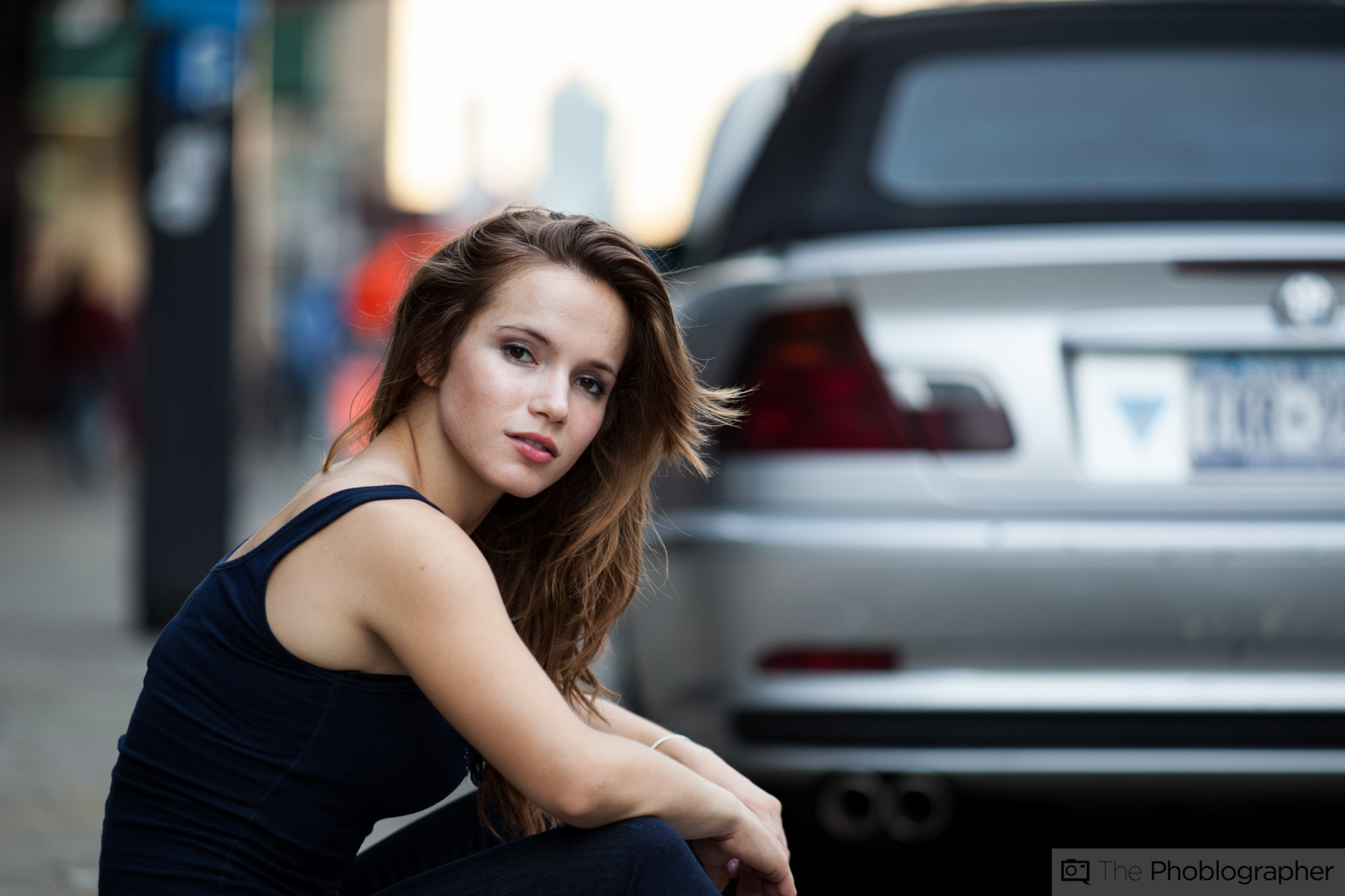 A young woman with long hair sits outdoors by a silver car, looking at the camera with a calm expression.