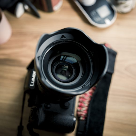 A close-up of a camera lens on a wooden desk with blurred objects in the background.