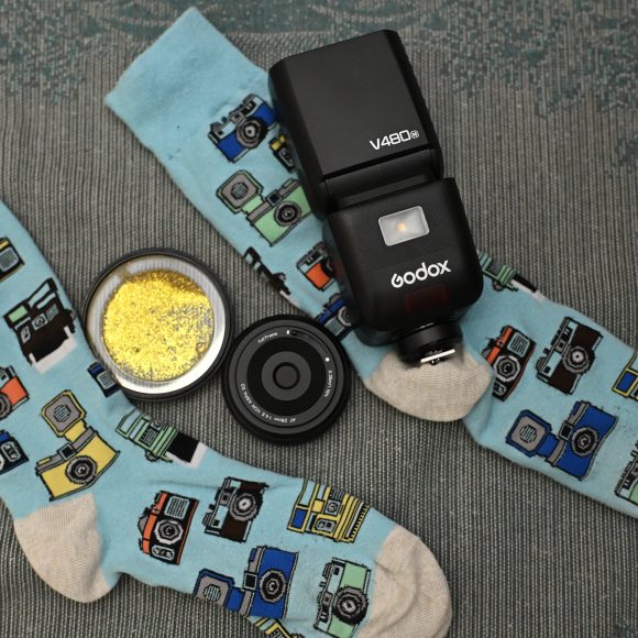 Stocking Stuffers For The Photographer In Your Life Two blue socks with camera prints, a camera flash, a camera lens, and a tin with yellow glitter.