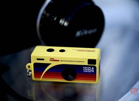 A small yellow toy camera keychain with 1984 and colorful stripes, next to a Nikon camera lens.