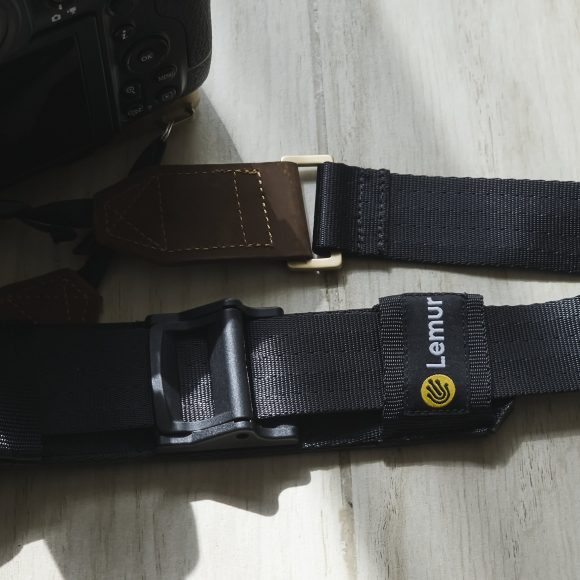 Lemur Strap: A Versatile Camera Strap That Really Sticks To You Close-up of a black Lemur camera strap with logo attached to a camera on a light wooden surface.