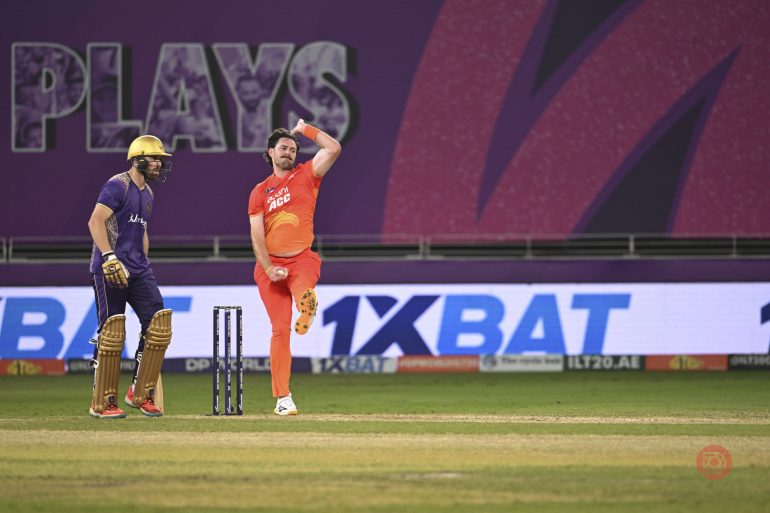 A cricket bowler in orange bowls as a batter in purple waits at the crease on a stadium field.