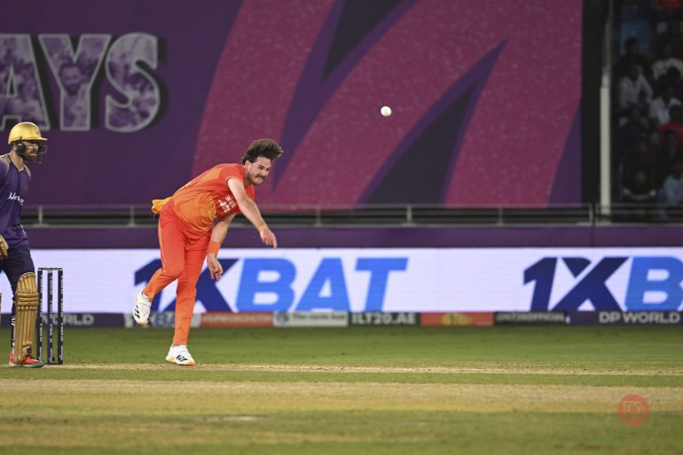 A bowler in an orange uniform delivers a ball during a cricket match as a batter waits at the crease.