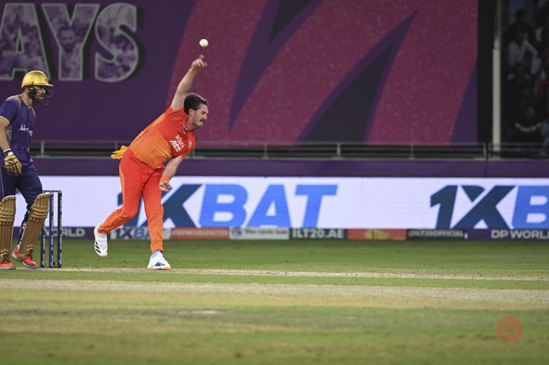 A cricket bowler in an orange uniform delivers the ball as a batsman and wicketkeeper look on.