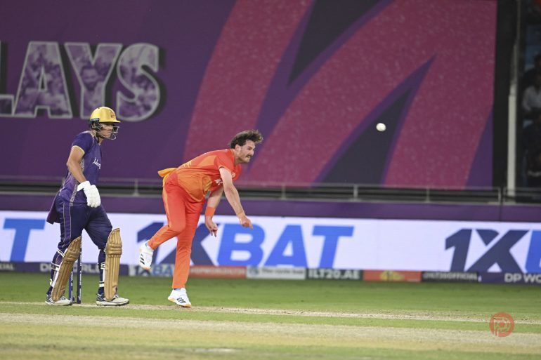 A bowler in orange delivers the ball as a batsman in purple watches during a cricket match.