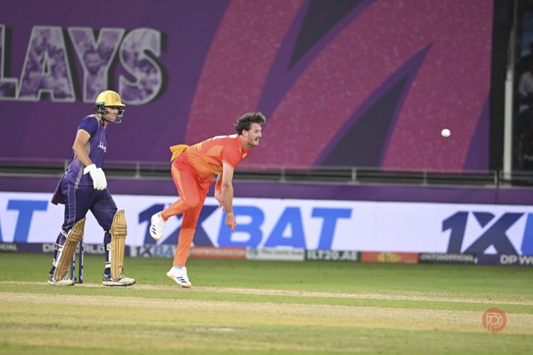 A bowler in orange mid-action delivers the ball as a batter in purple watches at the cricket crease.