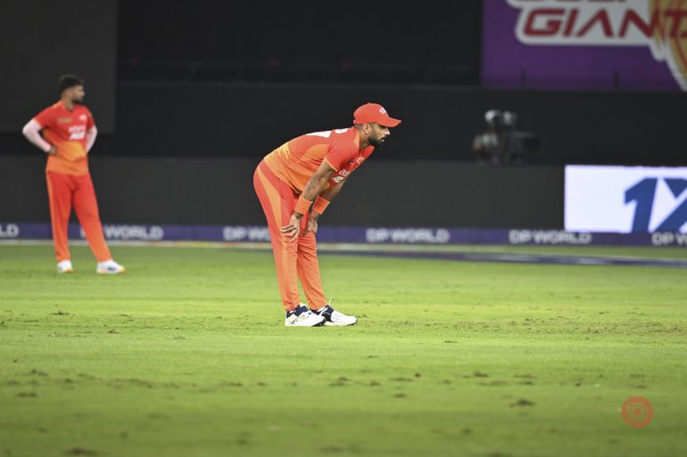 A cricketer in an orange uniform stands on a field, leaning forward with hands on knees.
