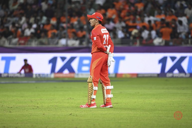 A cricket player in red gear stands on the field during a match, with a crowd in the background.