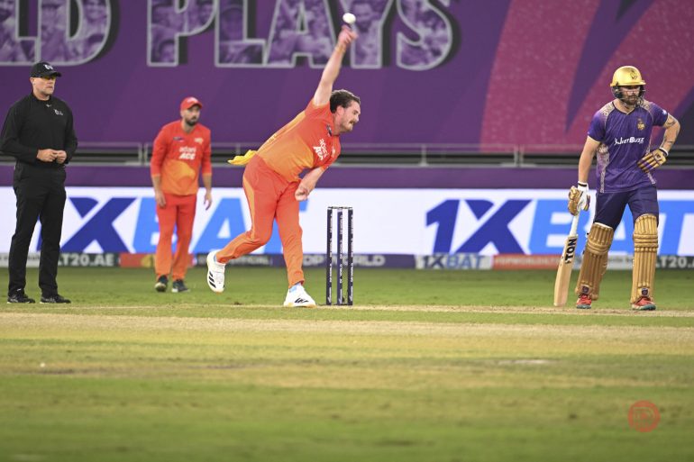A cricket bowler in orange delivers the ball as a batter in purple prepares to face it, with umpire watching.