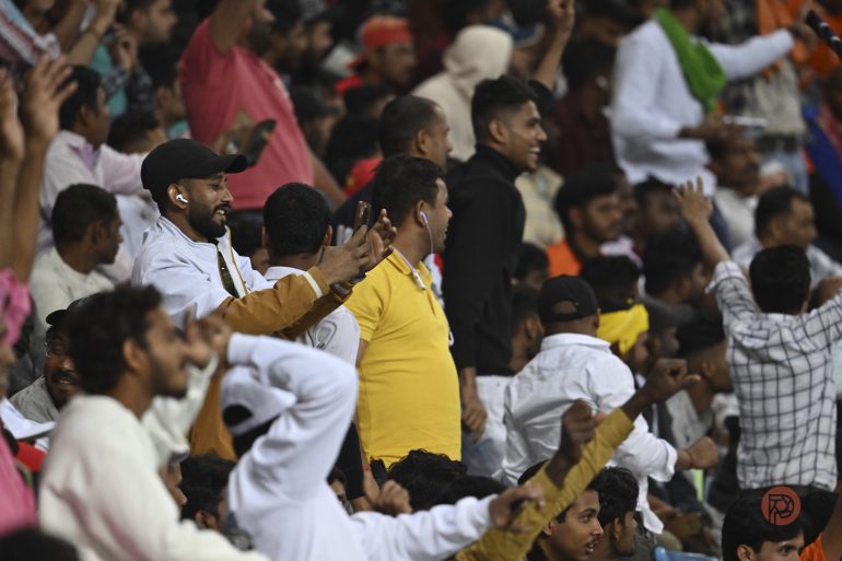 Cheering crowd at a stadium, some fans capturing the moment on their phones, all dressed in casual clothes.