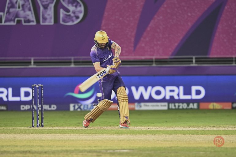 Cricketer in purple helmet bats during a match, with stumps and ball visible on the field.