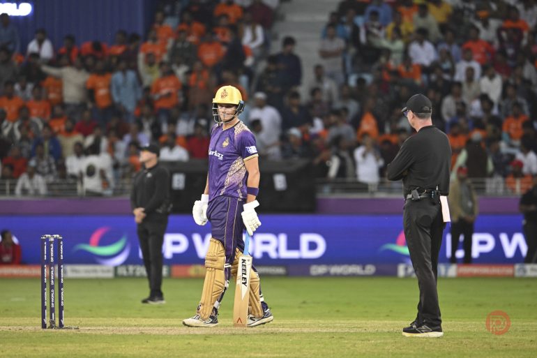 A cricket player in purple walks near the stumps as an umpire stands nearby; crowd in the background.