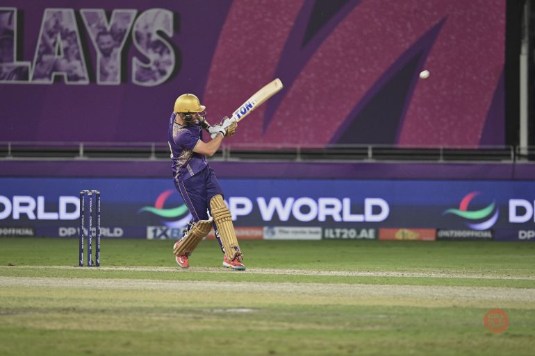 Cricket player in purple and gold hits the ball mid-air during a match on a lit stadium field.