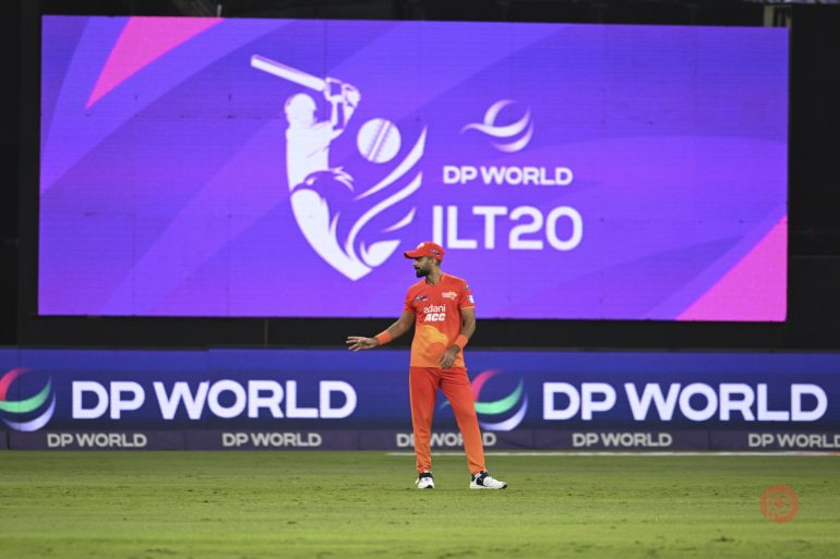 A cricket player in an orange uniform stands on a field with ILT20 and DP World logos in the background.