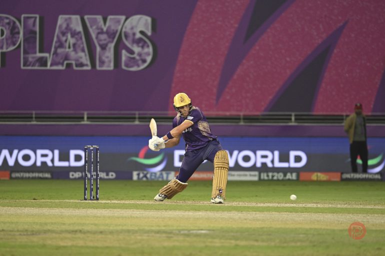 A cricketer in purple and yellow gear plays a shot during a match on a stadium field.