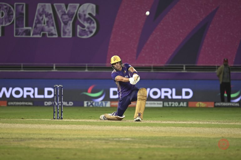 Cricketer in purple uniform plays a shot on a green field during a match, with stumps and ball visible.