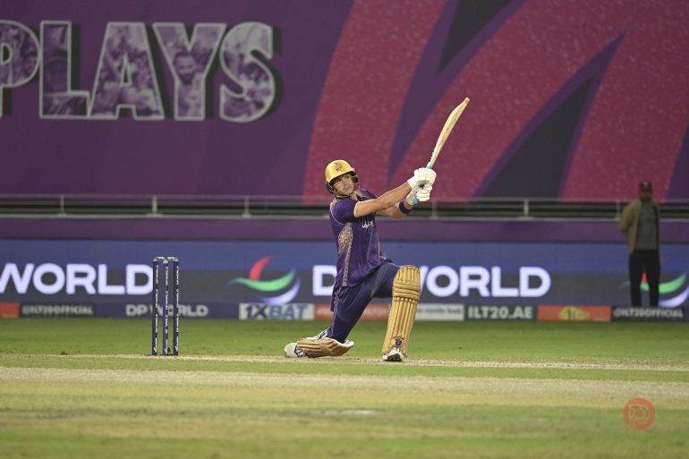 A cricket player in purple hits a shot on one knee during a match, with stumps and purple signage behind.