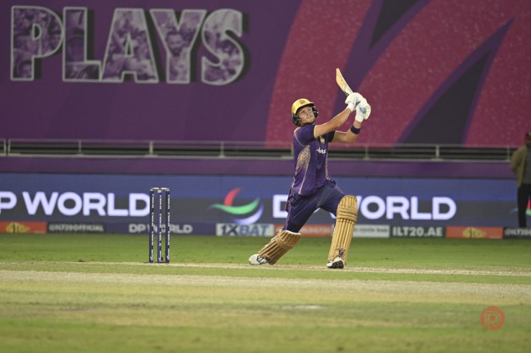 A cricket player in purple hits a shot, looking up at the ball, in a stadium with purple signage.