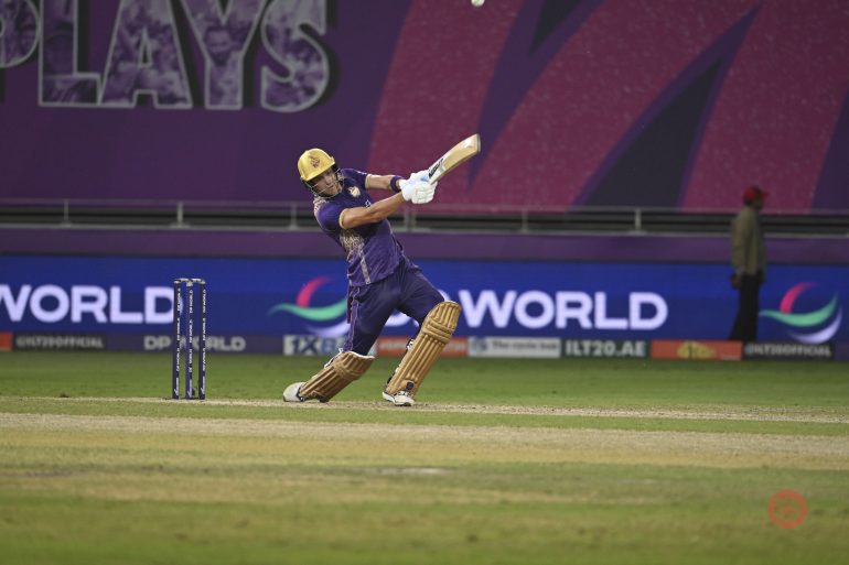 Cricketer in purple kit bats during a match on a green field with a purple stadium background.