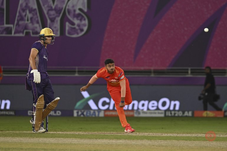 A cricket bowler in orange delivers the ball as a batter in gold and purple waits at the crease.