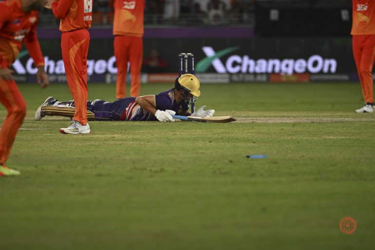 A cricket player dives to reach the crease as fielders in orange uniforms stand nearby on the field.
