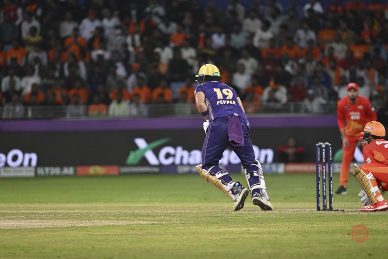 A cricket batter in purple and yellow gear runs between wickets during a match with a crowd in the background.