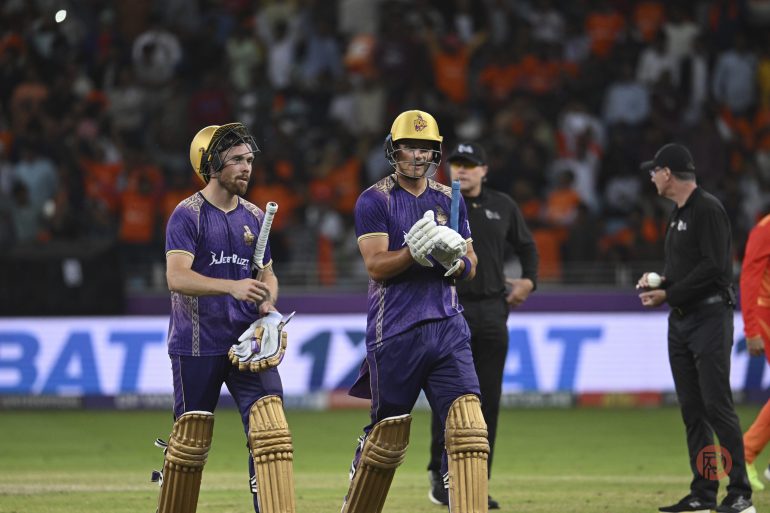Two cricket players in purple jerseys and helmets walk off the field holding bats, with a crowd in the background.